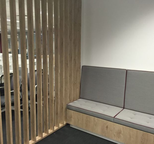 A compact contemporary office or commercial breakout area with a built-in upholstered bench seat beside a vertical timber slat privacy screen. The bench has grey cushions with dark red piping and a timber-look base, set against a plain white wall and dark carpet. Office furniture is partially visible through the slatted divider.