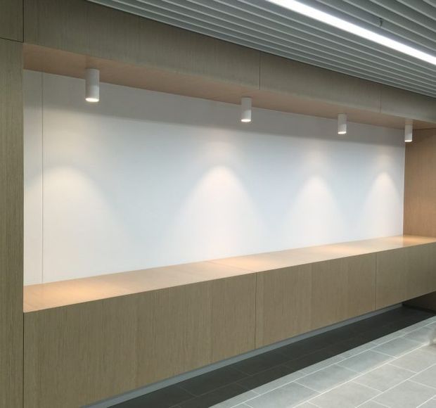 Minimalist commercial interior featuring a long built-in timber-look reception or display counter against a white wall, illuminated by evenly spaced cylindrical downlights. The space has a linear ribbed metal ceiling, light wood wall and joinery finishes, and dark tiled flooring.