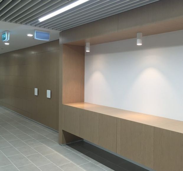 A clean, modern commercial interior corridor or lobby with a long built-in timber-look bench and wall cabinetry, a white illuminated feature wall, grey tiled flooring, recessed and pendant-style lighting, and a slatted linear ceiling. The space appears functional and minimalist, with no loose furnishings or people visible.