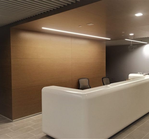 A refined commercial reception area featuring a large curved white reception desk, warm timber wall panelling, grey tiled flooring, dark grey walls, and integrated linear and recessed lighting. A slatted ceiling element adds texture and visual interest.