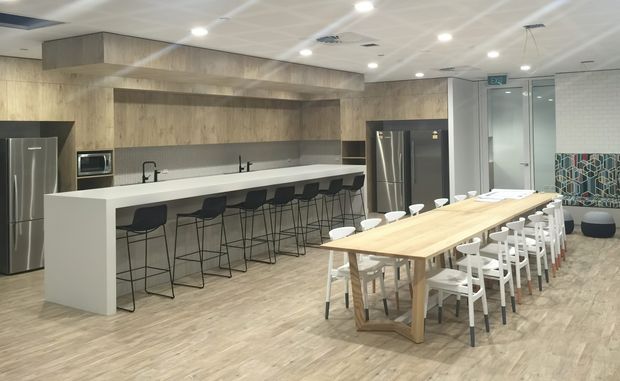 A spacious contemporary communal kitchen and dining area with light timber-look wall panelling and flooring, a long white island with black bar stools, stainless-steel refrigerators and appliances, and a large timber dining table surrounded by mixed white and black seating. Recessed ceiling lighting and geometric patterned tilework add visual interest.