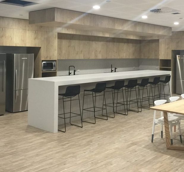 A spacious contemporary kitchen and communal dining area featuring a long white island with integrated sinks and seating for several people. The kitchen has extensive light timber cabinetry and wall panelling, a grey textured backsplash, stainless-steel refrigerators, integrated appliances, black tapware, and a light timber-look floor. A timber dining table with white chairs is partially visible in the foreground.