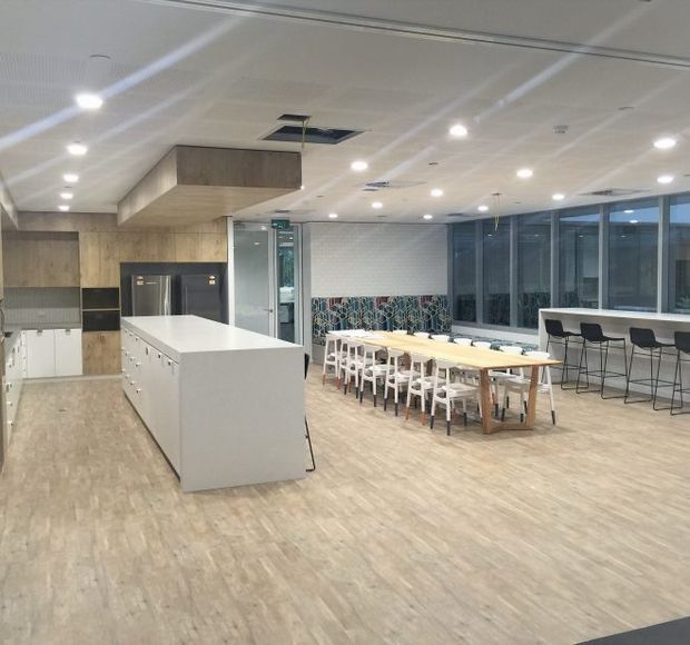 Bright contemporary communal kitchen and dining area with a large white island, integrated timber cabinetry, a long shared dining table, bar seating, expansive windows, and light timber-look flooring. The space appears designed for collaborative workplace or commercial use.