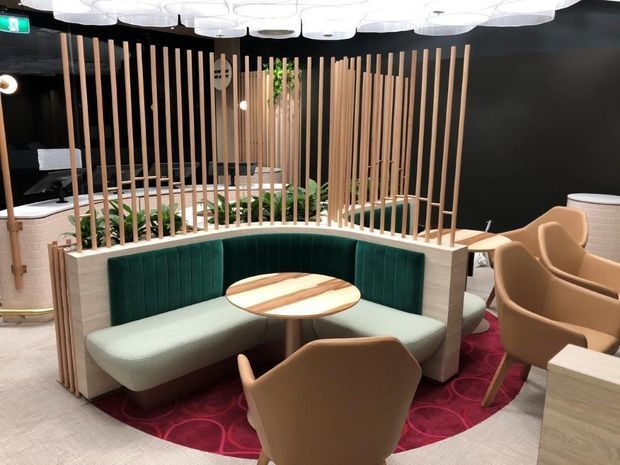 A refined commercial hospitality or lounge interior featuring a curved upholstered banquette in deep green, a round timber-topped table, and surrounding tan armchairs. Vertical timber slats form a semi-enclosed screen around the seating, with a black feature wall and sculptural white ceiling lighting. A patterned red rug and neutral carpet add contrast, creating an intimate, contemporary space.