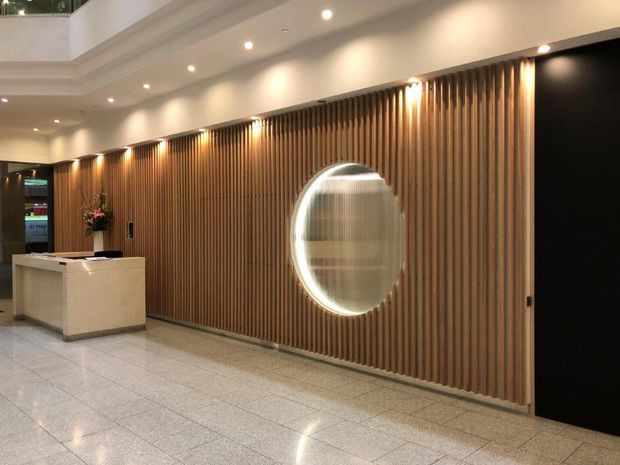 A refined commercial lobby or reception area with a white stone reception desk, a full-height vertical timber slat feature wall, a circular illuminated wall opening or mirror, recessed ceiling lighting, and a polished light terrazzo or stone floor. The space has a warm, contemporary hospitality or office fit-out.