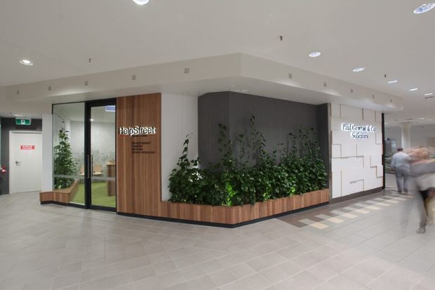 A contemporary commercial interior lobby or corridor featuring a branded HelpStreet office entrance. The space has pale tiled flooring, a white suspended ceiling with recessed circular lights, glazed office frontage, warm vertical timber cladding around the entry, and a large built-in planter with lush greenery. A white geometric wall with directional or tenant signage is visible to the right, with people passing through the corridor.