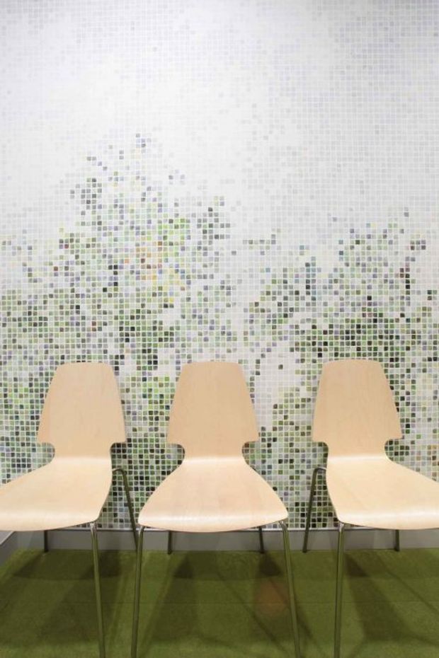 A bright interior featuring three light timber sculptural chairs arranged in a row against a full-height pixelated mosaic feature wall in soft white, grey, green and muted yellow tones. The chairs stand on a green carpet, creating a clean, playful and contemporary waiting or seating area.