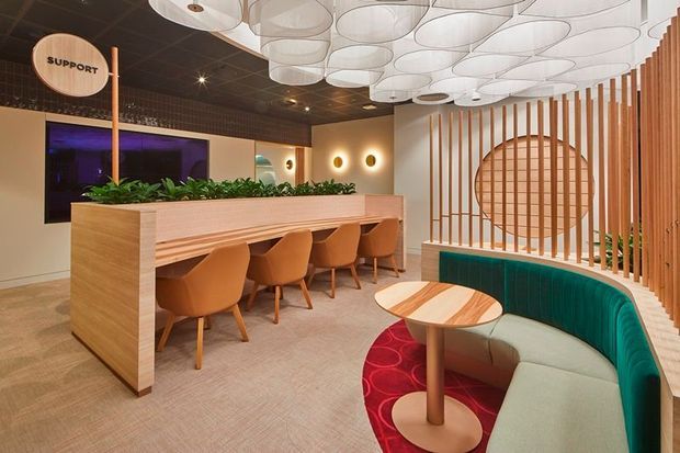A contemporary commercial hospitality or workplace lounge with a built-in timber communal counter, upholstered dining chairs, curved banquette seating, a round timber table, indoor planting, and a decorative vertical timber screen. The space features a dark gridded ceiling with layered circular pendant lights, warm ambient wall lighting, and a large display screen.