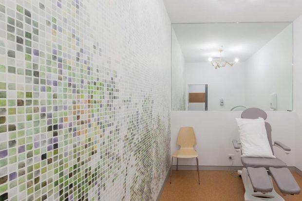 Bright, clinical treatment or consultation room with a large wall of small square mosaic tiles in white, pale green, lavender and muted blue. A reclining examination chair occupies the foreground, with a simple timber chair, wall-mounted fixtures, a large mirror, and a small chandelier-like ceiling light. The space has white walls, a warm tan floor, and a clean minimalist aesthetic.
