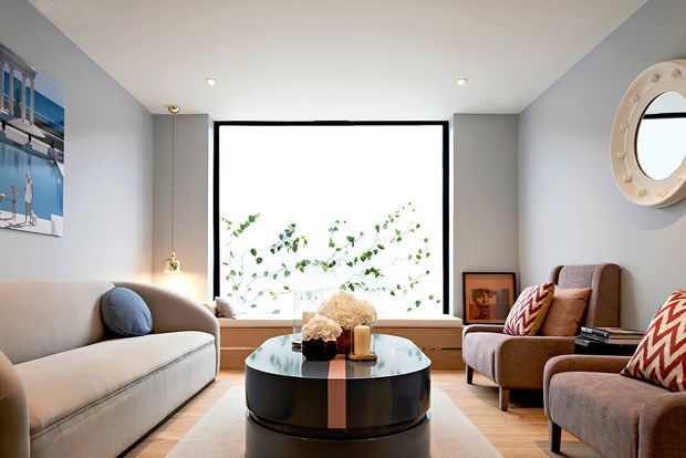 A refined living room with a large black-framed picture window, pale grey walls, a light upholstered sofa, two brown armchairs, and a glossy black round coffee table. Decorative cushions, artwork, a porthole-style wall mirror, and indoor plants add character to the bright, balanced space.