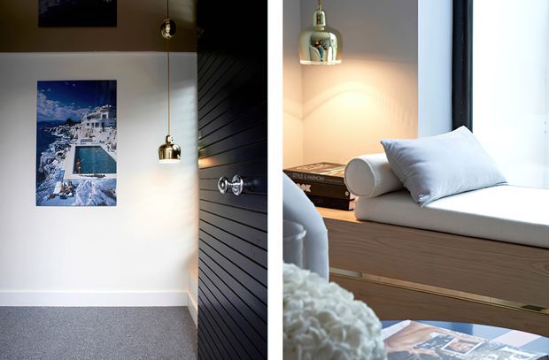 A split interior view showing a refined contemporary room. On the left, a white wall with framed artwork, pendant lights, grey carpet, and a dark vertical-slat door or wall panel. On the right, a bright window-side built-in window seat with white upholstery and cushions, pale timber cabinetry, books, and metallic wall lighting. The space has a clean, warm, modern feel with strong natural light.