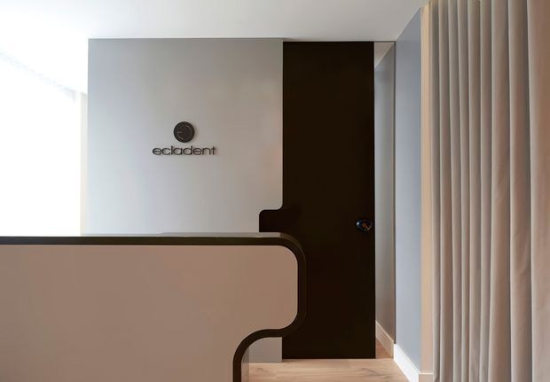 A refined commercial interior reception or waiting area with a curved upholstered partition or desk in the foreground, a white branded wall, dark timber-toned door, pale timber flooring, and full-height vertical curtains along the right side. The space has a restrained, contemporary fit-out with soft neutral tones and clean geometric lines.
