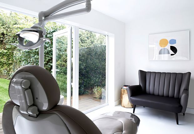 Bright, minimalist dental treatment room with a dental chair and overhead examination light in the foreground. Full-height glazed doors open to a small timber deck and leafy garden, while a sculptural black upholstered sofa and abstract artwork add a refined waiting-lounge feel.