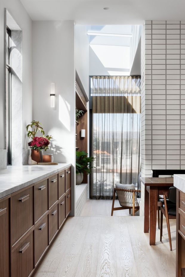 Bright, narrow contemporary interior combining a kitchen and dining area around a tall light-filled void. The space features a long run of dark timber cabinetry with metal pulls, pale timber flooring, white stone-like benchtops, a white horizontally tiled feature wall, a skylight, sheer curtains, and a large glazed opening. A small dining table and woven chair sit at the far end, with indoor plants and sculptural decor adding warmth.