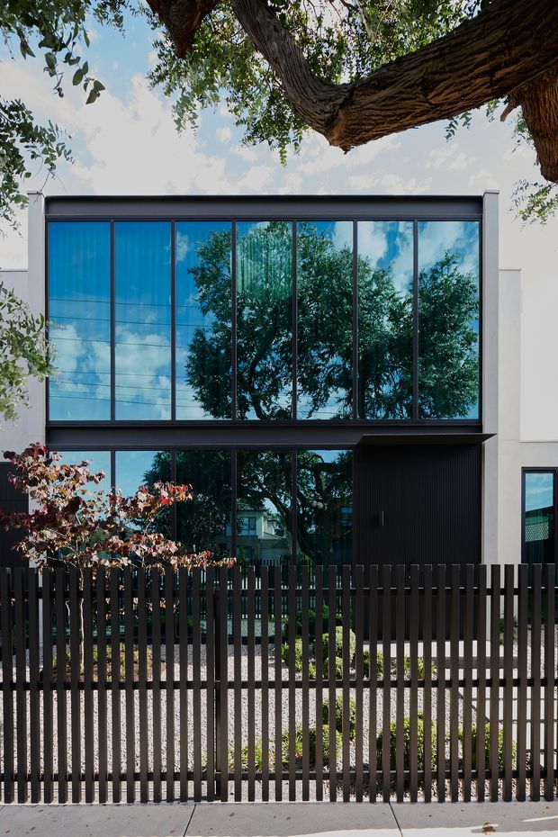 A contemporary two-storey residential facade viewed from the street, featuring extensive floor-to-ceiling dark-framed glazing, white rendered wall panels, a dark vertically slatted entry or garage screen, and a matching vertical-slat front fence. Mature tree branches frame the upper portion, while reflections of surrounding trees and sky appear in the glazing.