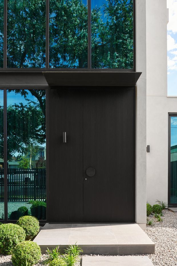 Contemporary residential facade detail featuring a tall, dark vertically fluted front door with a minimalist handle and circular hardware. Large dark-framed glazed panels reflect mature trees, while pale rendered walls, a tiled entry landing, and gravel landscaping with clipped shrubs complete the composition.