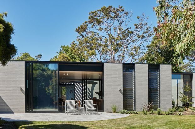 Single-storey contemporary residence with a low, flat-roofed profile, dark-framed glazing, and light grey masonry facade panels. Large glazed openings connect the interior to a paved outdoor sitting area, with a neatly landscaped lawn and established trees surrounding the home.