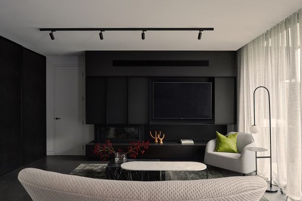 A sophisticated modern living room with a dark built-in media wall and recessed television, black cabinetry, and a low linear fireplace or display niche. A curved textured sofa faces the media unit, accompanied by a sculptural armchair, small side table, marble-like oval coffee table, and patterned rug. Full-height sheer curtains provide soft daylight along the glazed wall, while black track lighting and a slim arched floor lamp add graphic accents.