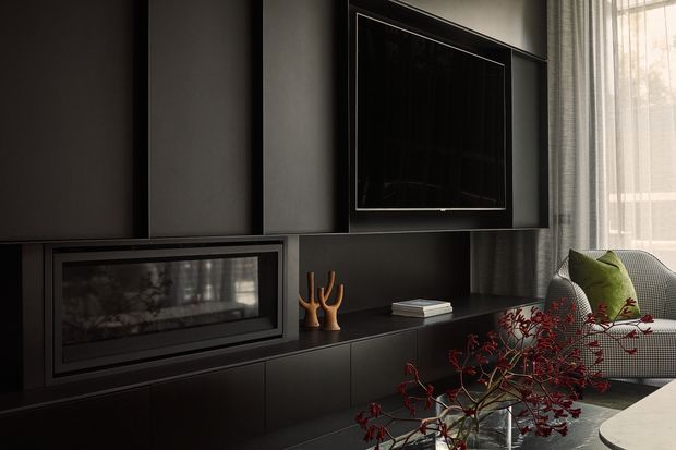 A moody contemporary living room media wall in dark charcoal tones, featuring a built-in linear fireplace, flush-mounted television, and extensive low cabinetry. A textured lounge chair with green cushions sits beside sheer floor-to-ceiling curtains, while sculptural timber objects, books, and red botanical branches add restrained accents.