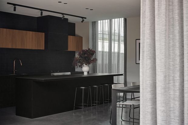 A dark, sophisticated open-plan kitchen and dining area. Matte black cabinetry and a black island with integrated seating dominate the room, contrasted by warm timber cabinet fronts. A black track-lighting system runs across the ceiling, while sheer floor-to-ceiling curtains frame a tall window. A compact dining table with dark metal-framed chairs is visible at right.