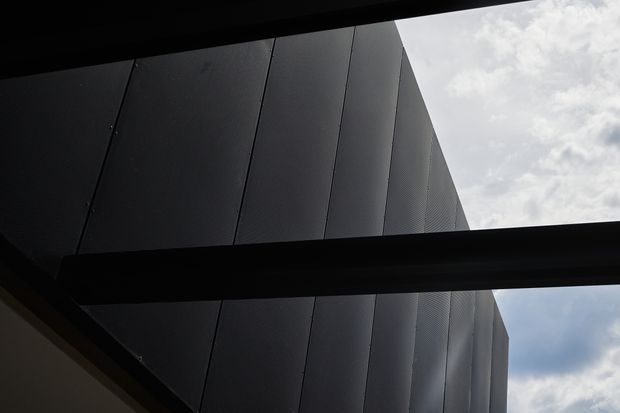 Upward-looking architectural detail showing a dark, angular building facade clad in large vertical panels, framed by heavy black structural beams or overhangs. A narrow opening reveals a partly cloudy sky, creating a stark, high-contrast composition with an industrial and contemporary character.