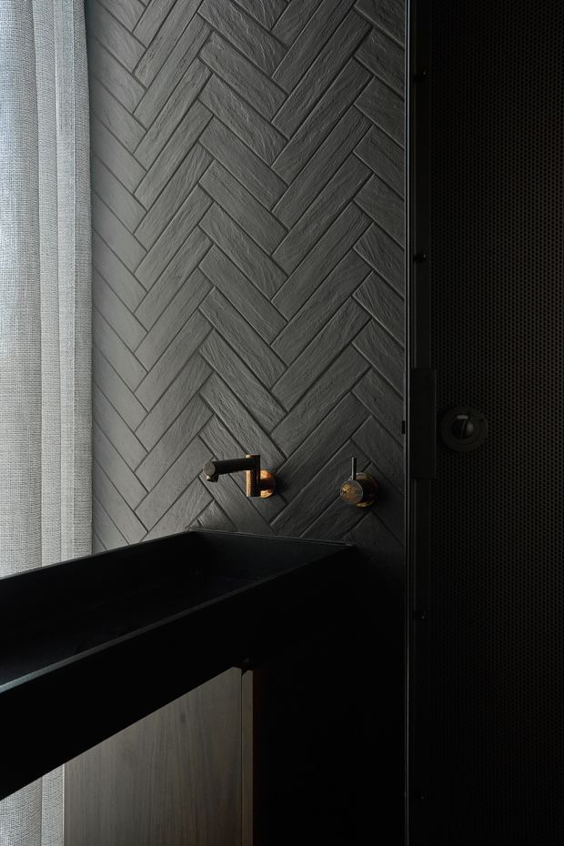 Close-up of a dark, moody bathroom featuring a charcoal grey herringbone wall tile installation, black freestanding or built-in bath edge in the foreground, and dark metal fixtures with warm brass accents. A sheer textured window covering provides soft filtered light at the left, while a black perforated or textured vertical panel frames the right side.