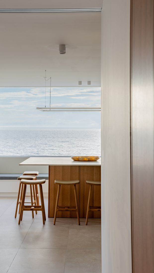 A minimalist kitchen and breakfast bar overlooking the ocean through expansive floor-to-ceiling glazing. The space features warm timber cabinetry and stools, a pale stone island benchtop, large-format light flooring, recessed ceiling details, and a slim linear pendant light. The composition is bright and serene, with a contemporary coastal character.