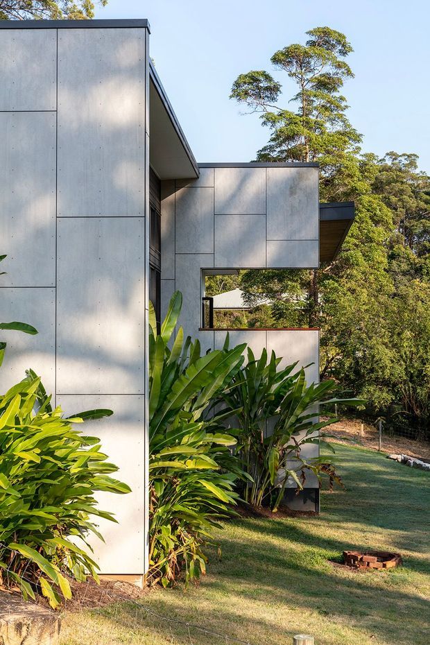 Exterior view of a contemporary, rectilinear house set within lush landscaped grounds. The building features large pale grey concrete panel cladding with visible joint lines, projecting box-like volumes, deep roof overhangs, dark-framed openings, and a partially visible timber-lined soffit. Tropical foliage and mature trees surround the structure.