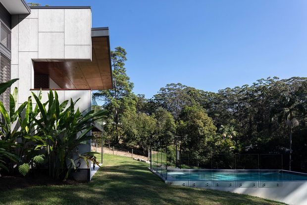 Exterior view of a modern multi-level house beside a swimming pool, set within a lush wooded landscape. The building features crisp pale panelled walls, dark-framed glazing, and a deep projecting upper-level canopy with a warm timber soffit. A lawn, tropical planting, pool fencing, and dense trees surround the home under a clear blue sky.
