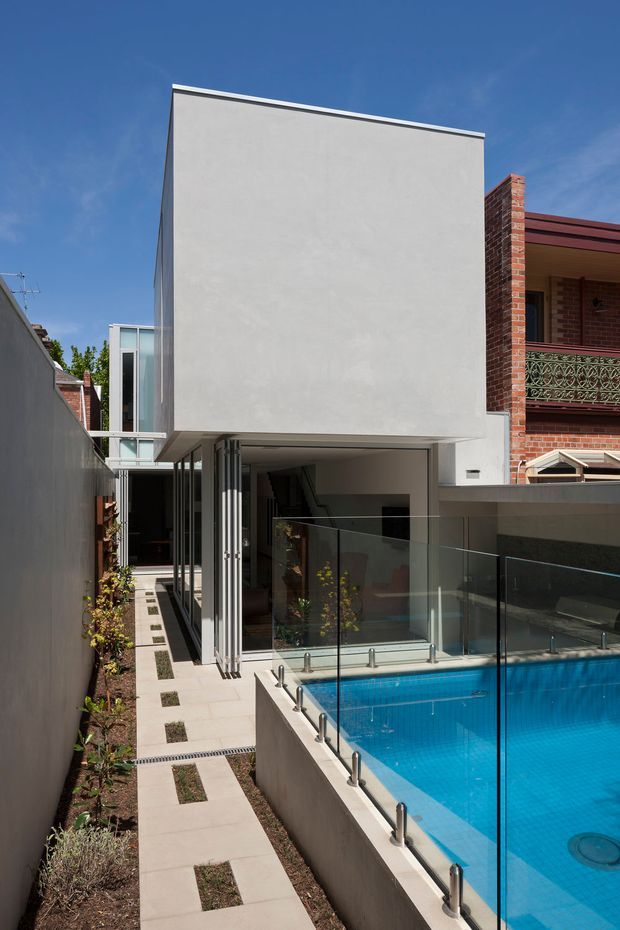 Exterior view of a narrow contemporary urban residence with a stark white rendered upper volume cantilevered over a glazed ground floor. A vivid blue swimming pool occupies the foreground, enclosed by frameless glass balustrades, with a paved side walkway and planted strip along the boundary. The neighbouring red-brick house remains visible on the right.