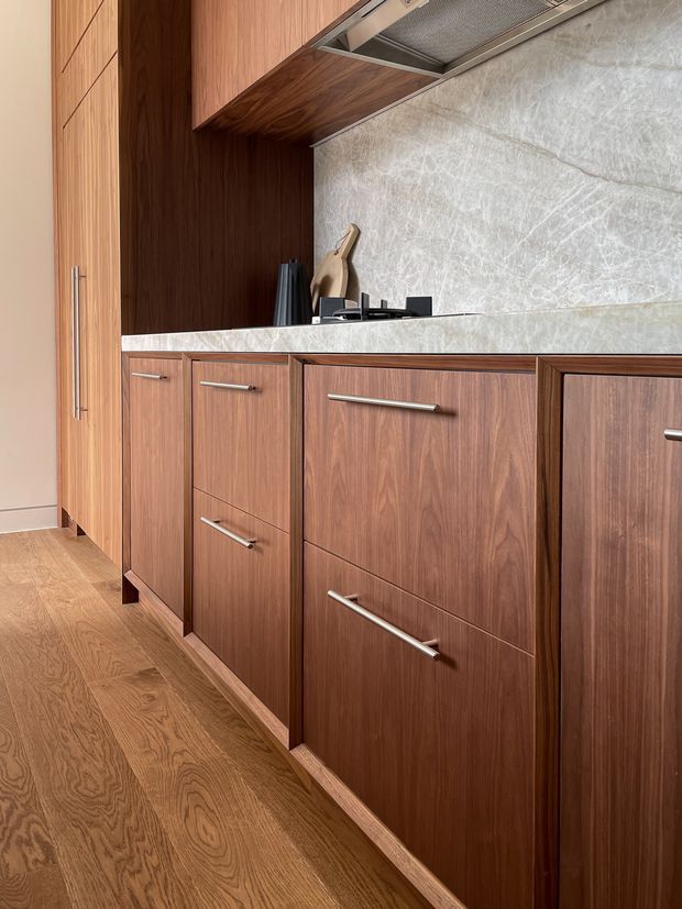 Close-up view of a refined kitchen joinery installation featuring dark walnut-toned wood cabinetry with long brushed-metal handles, a pale veined stone benchtop and splashback, an integrated stainless-steel rangehood, and warm timber flooring. The cabinetry includes tall pantry-style storage and multiple drawers, creating a streamlined contemporary kitchen composition.