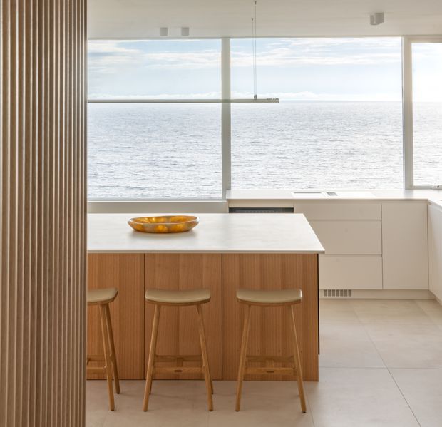 A bright, minimalist kitchen overlooking the ocean through expansive floor-to-ceiling windows. The space features a white island benchtop with integrated seating, warm vertical timber-faced cabinetry, white perimeter cabinetry, pale tiled flooring, and a slim linear pendant light. The ocean view creates a strong coastal setting.