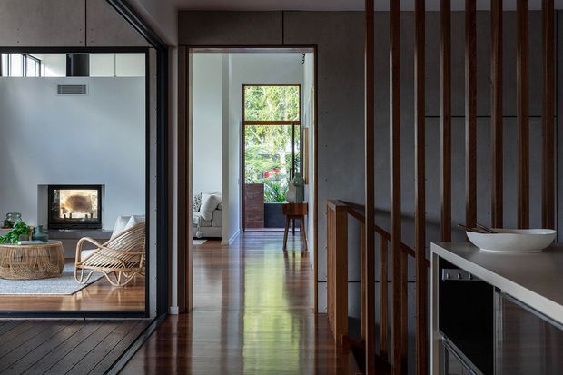 Interior view along a polished timber hallway, with a living room and fireplace visible through full-height glazing on the left. Vertical dark timber screens and a slim console with a white vessel basin define the right side, while a bright seating area and garden view are visible at the end of the corridor. The space has a refined contemporary character with strong linear detailing and warm natural materials.