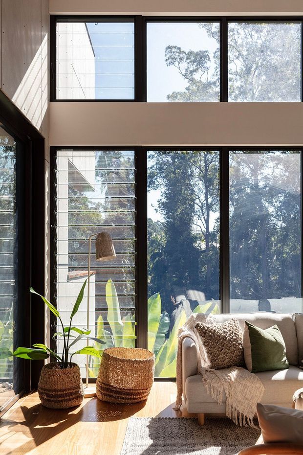 Sunlit living room with double-height glazing, dark-framed windows and louvred panels overlooking leafy trees. A neutral sofa with textured cushions and throw sits beside woven planters and a floor lamp on a warm timber floor, creating a bright, natural interior.