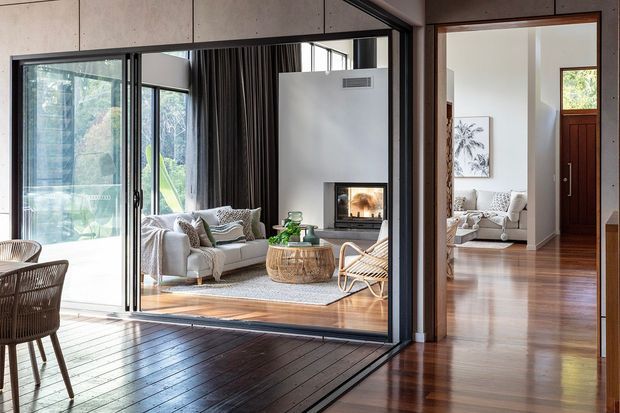 A bright contemporary residential interior viewed from a dark timber-decked dining area, looking through large black-framed glazed sliding doors into a living room. The living room features a pale sofa, textured rug, woven coffee table and chairs, dark curtains, a white fireplace wall with a built-in firebox, and extensive garden-facing glazing. A second sitting area with timber flooring is visible through an internal opening on the right.
