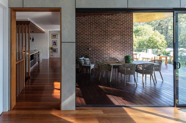 Interior view looking toward a dining area opening onto a timber deck and garden. A large dark-framed glazed opening, exposed red-brick feature wall, concrete panel walls, timber flooring, and timber slatted screening define the space. The adjacent hallway and stair railing are visible through the doorway on the left, while the outdoor dining setting is visible beyond the glazing.