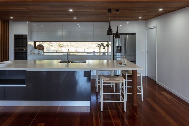 A contemporary open-plan kitchen dominated by a large island with a light stone benchtop and dark fixed cabinetry. Glossy white upper cabinetry spans the back wall, with integrated ovens, a stainless-steel refrigerator, sink, cooktop, and a long horizontal window providing natural light. The room features rich dark timber flooring, a timber-lined ceiling, white walls, and two dark pendant lights over the island, with stools tucked beneath the breakfast bar.