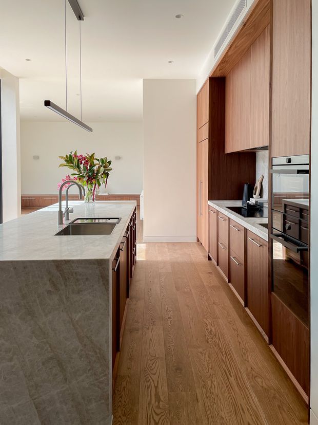 Contemporary galley-style kitchen with a large stone island, integrated sink and curved chrome tap, extensive warm timber cabinetry, built-in ovens, and a light timber floor. The space has white walls and ceiling, a linear suspended pendant, and a bright, refined material palette.