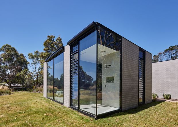 A compact contemporary black-brick pavilion set within a landscaped lawn, with extensive floor-to-ceiling glazing, dark framed windows, and external louvres. A bathroom with a shower is clearly visible through the transparent corner glazing, while trees and lawn surround the structure. The design has a crisp cubic form and a flat roof.