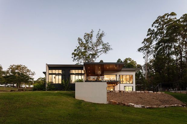 Exterior view of a contemporary two-storey residence set within a landscaped, semi-rural site. The house has a clean rectilinear form, extensive window walls, dark framed glazing, pale rendered wall surfaces, and a projecting upper-level roof or canopy over a sheltered entry and balcony area. A broad lawn, planted trees, gravel landscaping, and a low rendered garden wall surround the building, photographed in soft evening light.