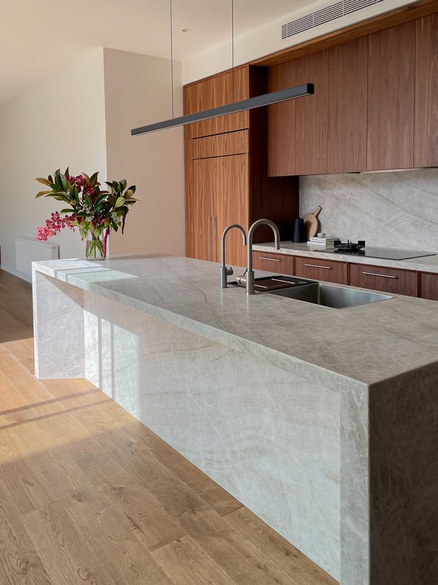 Contemporary kitchen interior dominated by a large pale stone island with an integrated double-bowl sink and tall curved tapware. Full-height dark timber cabinetry lines the rear wall, with matching lower cabinets, a pale veined stone splashback, and a concealed cooktop. Warm timber flooring, neutral walls, and a slim black linear pendant create a refined, minimalist atmosphere.