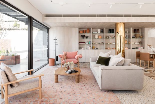 A bright, spacious contemporary living room with floor-to-ceiling glazing, a pale upholstered sectional sofa, timber-framed lounge chair, pink accent chair, low timber coffee table, and a large pastel patterned rug. Full-height light timber built-in shelving and cabinetry line the rear wall, displaying books and decorative objects, while a polished brass cylindrical column creates a strong feature. A dining area is visible beyond the living space.
