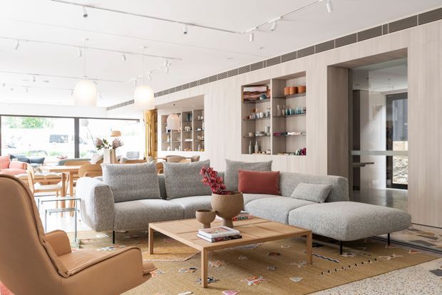 Bright, open-plan contemporary living room with a large L-shaped grey sectional sofa, tan lounge chairs, a timber coffee table, and built-in light-wood shelving displaying books and ceramics. The space includes a dining area and kitchen or service area in the background, expansive glazing, white ceilings with track lighting, and a warm patterned rug. The design is refined, airy, and understated, with integrated linear ventilation and extensive built-in joinery.