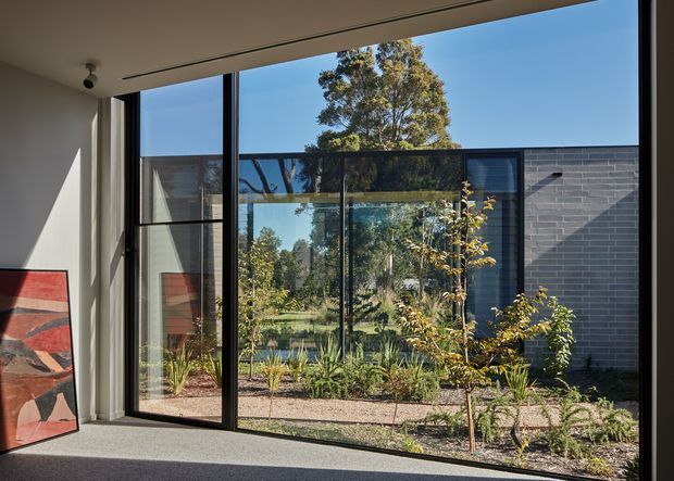 A contemporary interior looking through large black-framed glazed doors and windows onto a landscaped courtyard garden. The room has a light neutral wall and ceiling, grey carpet, and a colourful abstract artwork leaning against the wall. Strong indoor-outdoor connection, with a dark brick exterior wall, gravel path, planting, and mature trees visible beyond the glazing.
