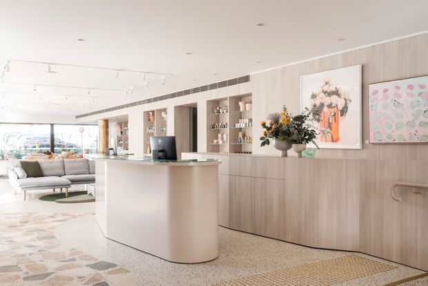 Bright contemporary open-plan interior featuring a curved reception or kitchen island with a pale stone benchtop, extensive light timber built-in cabinetry and shelving, integrated appliances, terrazzo-style flooring, artwork, and a lounge area beyond. Full-height glazing brings in abundant daylight, creating an airy, refined space.