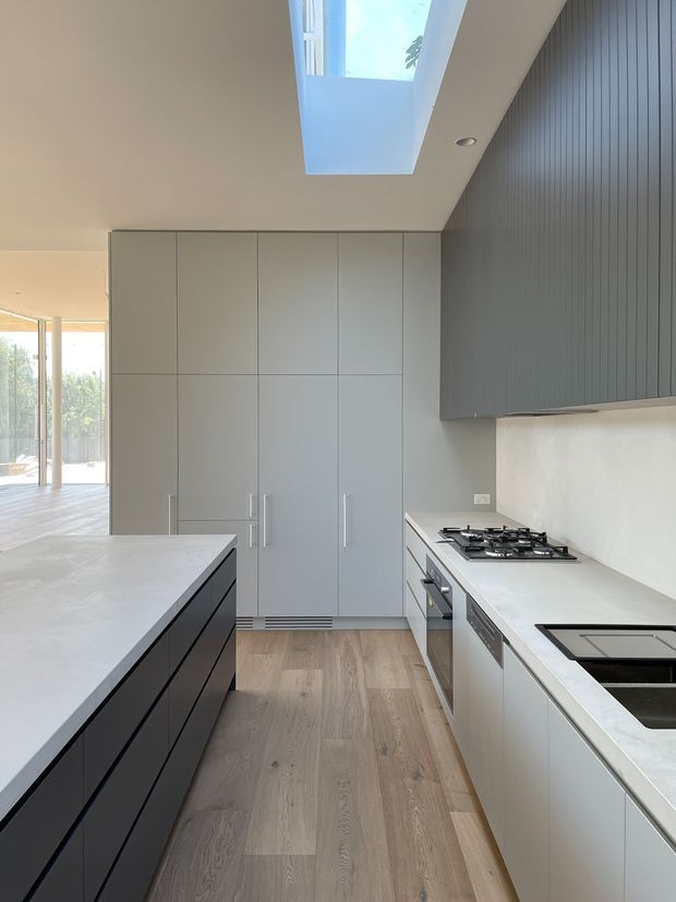 A bright contemporary galley kitchen with a large central island, pale stone benchtops, light grey full-height cabinetry, and dark vertically ribbed overhead cabinetry. Integrated appliances, a gas cooktop, and a black sink are visible. The kitchen has light timber flooring, a large skylight, and an open connection to a glazed living or outdoor area.