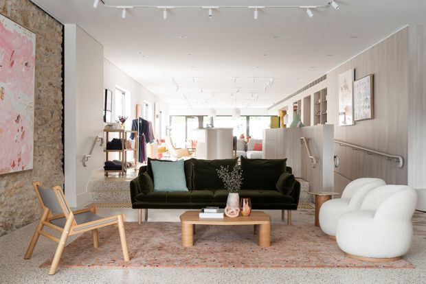 A bright, open-plan contemporary living room with a deep green velvet sofa, sculptural white lounge chairs, a timber coffee table, and a large patterned rug. The room features pale vertical wall panelling, built-in shelving, framed artwork, track lighting, a textured exposed-stone feature wall, and a visible dining or kitchen area beyond. A small set of steps and handrails connect the living area to the rear zone.