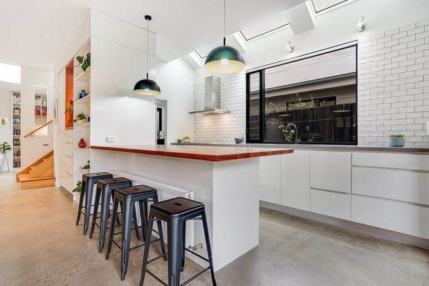 Bright contemporary kitchen with a long island and four black metal stools, white flat-panel cabinetry, white subway-tile splashback, and a warm timber island benchtop edge. Skylights and pendant lights provide abundant natural and task lighting. A hallway and staircase are visible beyond the kitchen.