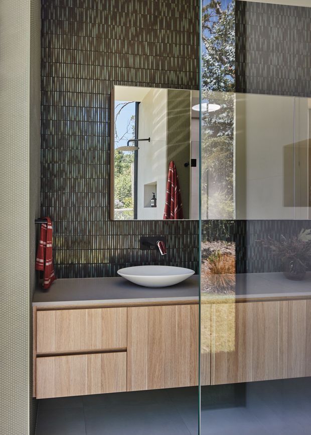 Contemporary bathroom vanity with a floating light-wood cabinet, dark benchtop, white vessel basin and wall-mounted tap. The feature wall is clad in narrow vertically patterned dark tiles, complemented by a large mirror and frameless glass panel. A red towel provides a small accent, while the mirror and glass reflect the adjoining shower and outdoor greenery.