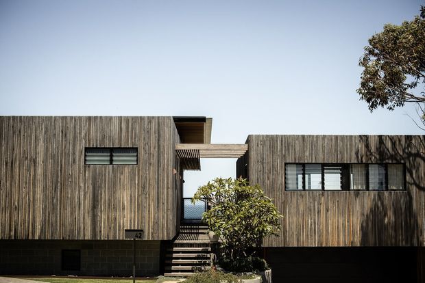 Front exterior view of a contemporary two-part dwelling composed of dark vertical timber-clad volumes connected by a raised covered bridge or entry canopy. Black masonry forms the lower level, with narrow horizontal windows, an external stair, and a central landscaped entry court. The composition has a crisp, minimalist geometric form under a clear sky.